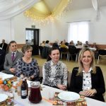 Grupa osób mężczyzn i kobiet siedzi przy świątecznym, wyłozonym białym obrusem stole i ogląda występ dzieci.