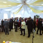 Grupa osób w jasnej sali widowiskowej dzieli się opłatkiem. Na suficie wiszą piekne, lniane dekoracje.