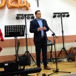 Młody mężczyzna w garniturze przemawia do mikrofonu na scenie, obok niego stoi starsza kobieta w czerwonej sukience.