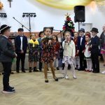 Występ dzieci ubrancyh w odświętne stroje na jasnej sali domu ludowego. Za dziećmi stoi pięknie przyozodobiona choinka.