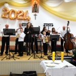 Występ kilkuosobowej grupy mężczyzn i kobiet na scenie z instrumentami muzycznymi.