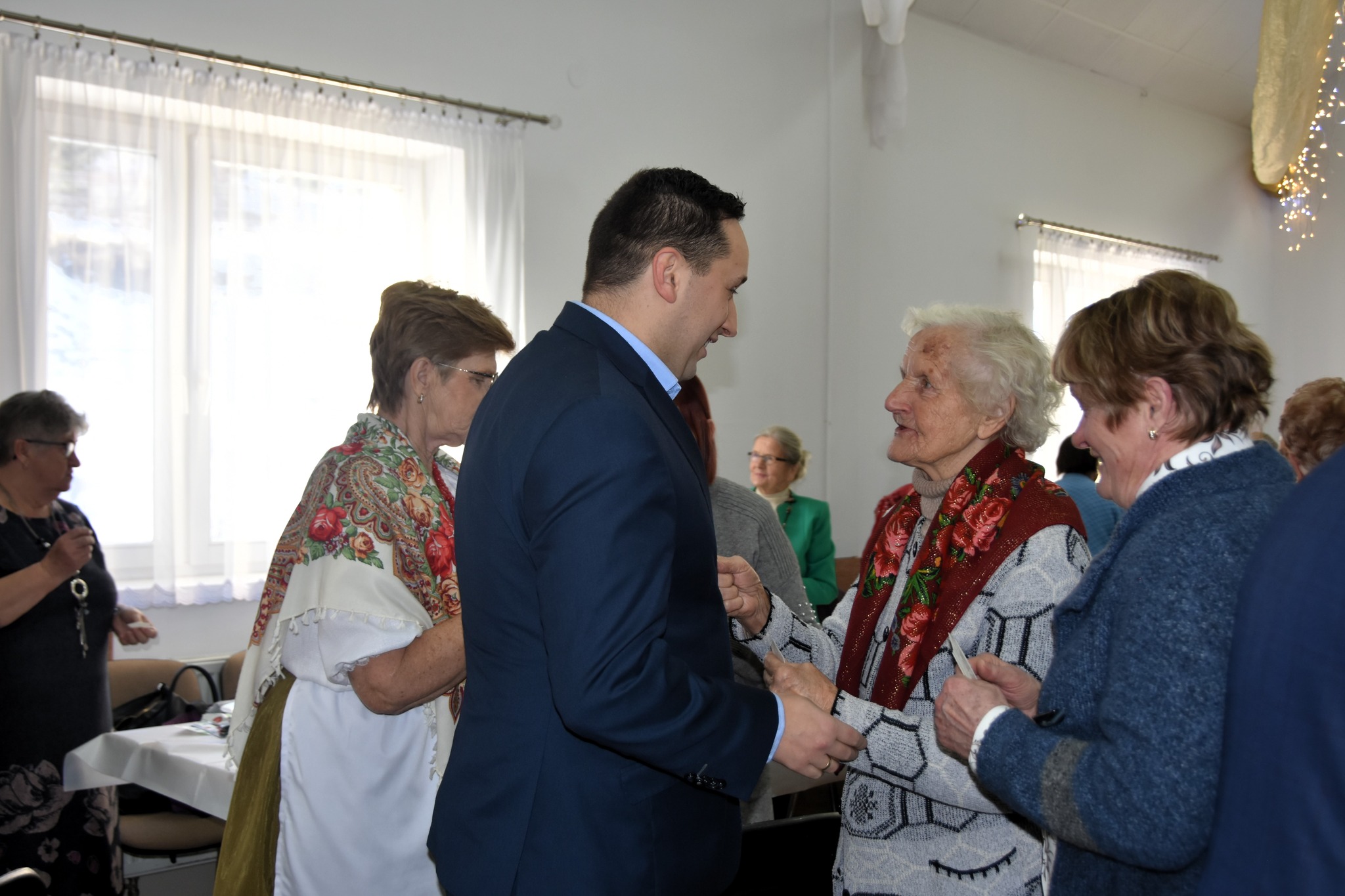 Młody mężczyzna w garniturze dzieli się oplatkiem ze starszą kobietą o siwych włosach, ubrana w swetr i zawieszoną na szyi chustę.