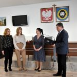 Mężczyzna w średnim wieku ubrany w garnitur stoi w jasnym pomieszczeniu z trzema kobietami. Dwie z nich ubrane są w spodnie i koszule, a trzecia w sukienkę. Za nimi na białej ścianie wiszą obrazy herbu gminy oraz herbu państwa. Obok wisi też telewizor.