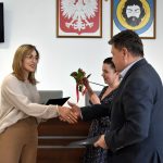 Męczyzna w garniturze przekazuje gratulacje, ściskając rękę kobiecie o blond włosach i przekazuje jej teczkę.