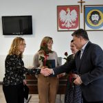 Męczyzna w garniturze przekazuje gratulacje, ściskając rękę kobiecie o blond włosach i przekazuje jej teczkę.