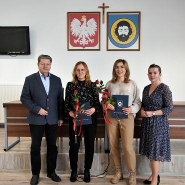 Mężczyzna w średnim wieku ubrany w garnitur stoi w jasnym pomieszczeniu z trzema kobietami. Dwie z nich ubrane są w spodnie i koszule, a trzecia w sukienkę. Za nimi na białej ścianie wiszą obrazy herbu gminy oraz herbu państwa. Obok wisi też telewizor.