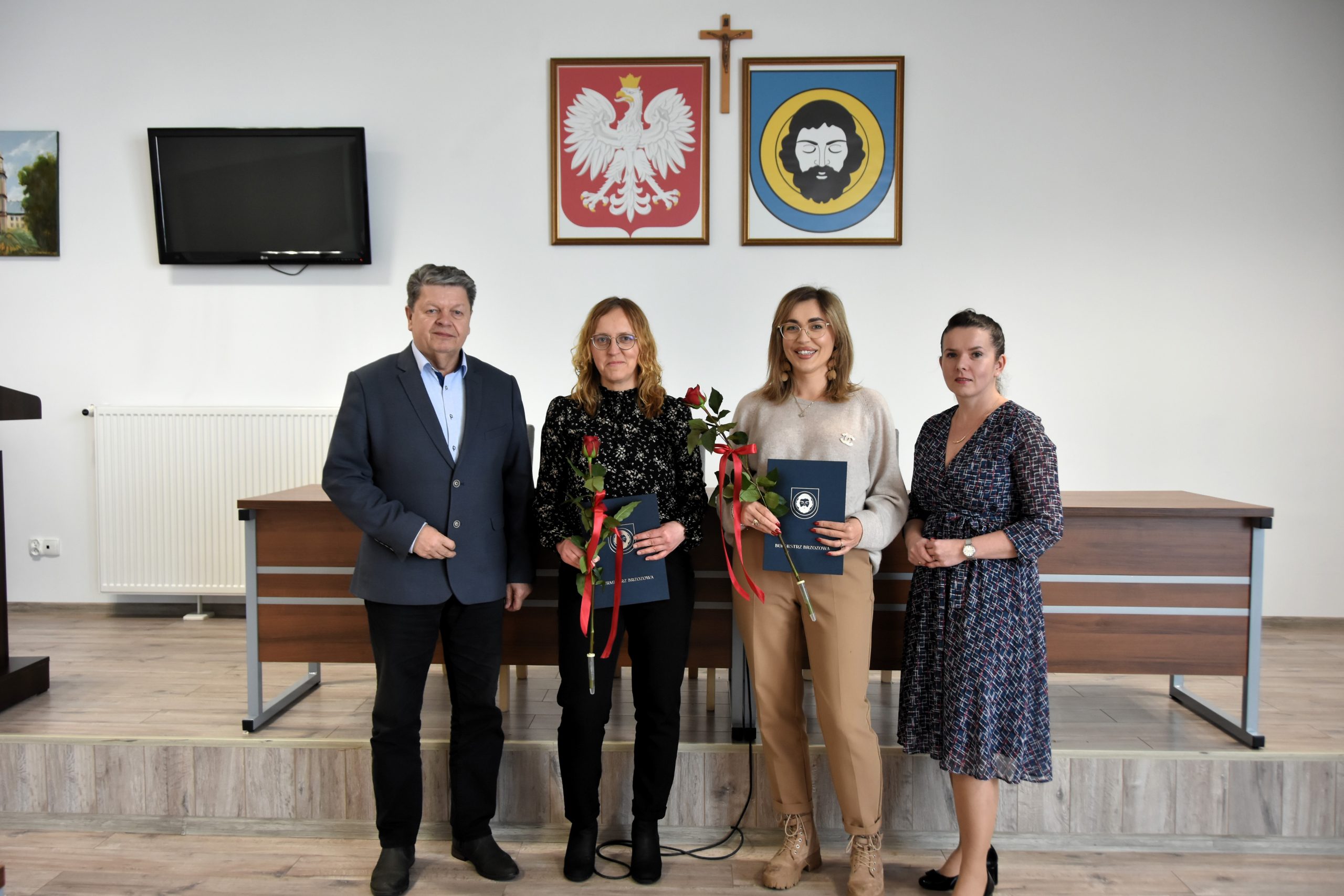 Mężczyzna w średnim wieku ubrany w garnitur stoi w jasnym pomieszczeniu z trzema kobietami. Dwie z nich ubrane są w spodnie i koszule, a trzecia w sukienkę. Za nimi na białej ścianie wiszą obrazy herbu gminy oraz herbu państwa. Obok wisi też telewizor.