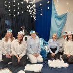 Parę kobiet w średnim wieku ubrane w koszulki białego i niebieskiego koloru oraz ciemne spodnie siedzą na podłodze w sali, za nimi dekracja kolorze ciemnym.