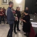 Dwóch mężczzyn składa na ręce straszej kobiety podziękowanie, wręczając jej bukiet kwiatów i torebkę prezentową.