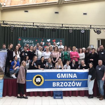 Grupa osób stoi na scenie, trzymając w rękach baner z napiesem Gmina Brzozów i pozuje do grupowego zdjęcia.