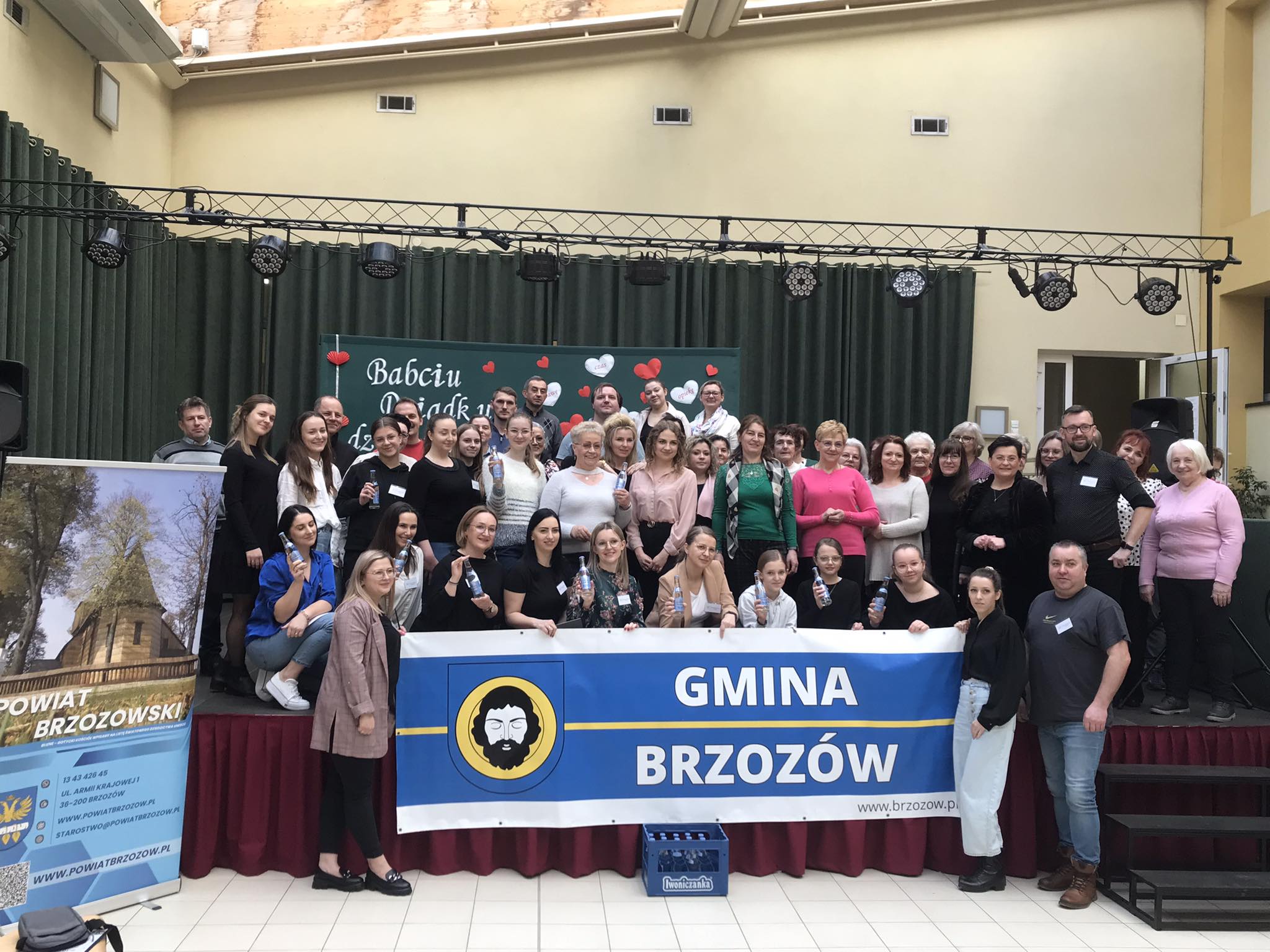 Grupa osób stoi na scenie, trzymając w rękach baner z napiesem Gmina Brzozów i pozuje do grupowego zdjęcia.