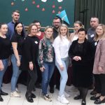 GHrupa osób stoi w sali i pozuje do zdjęcia, za nimi zielone tło.