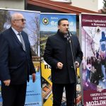 Młody mężczyzna w garniturze przemawia do mikrofonu na zewnątrz, za nim stoją ustawione banery promocyjne.