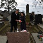 Starszy mężczyzna w garniturze i młoda kobieta stoją przy grobie na cmentarzu i odpalają znicz.