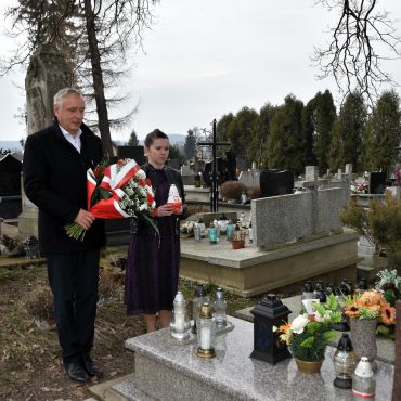 Starszy mężczyzna w garniturze i młoda kobieta stoją przy grobie na cmentarzu. Mężczyzna trzyma w rękach wiązankę kwiatów, zaś kobieta znicz.