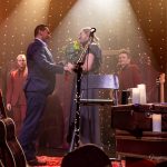 Młody mężczyzna w garniturze wręcza kobiecie o blond włosach i w szarej sukience, bukiet czerwonych róż. Za nimi na scenie stoją męźczyżni w czerownych garniturach.