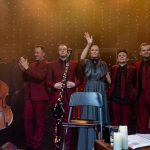 Grupa 5 osób, czterech mężczyzn i kobieta w środku stoją na scenie i machają do publiczności.