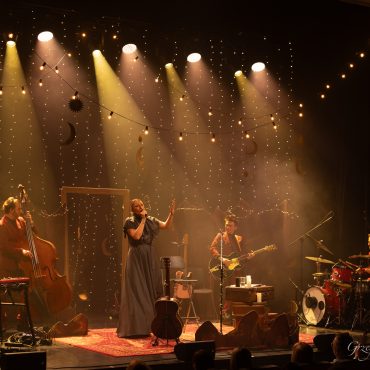 Kobieta o blond włosach siedzi na krześle i spiewa do mikrofonu, za nią na instrumentach grają męźczyżni w czerwonych garniturach.