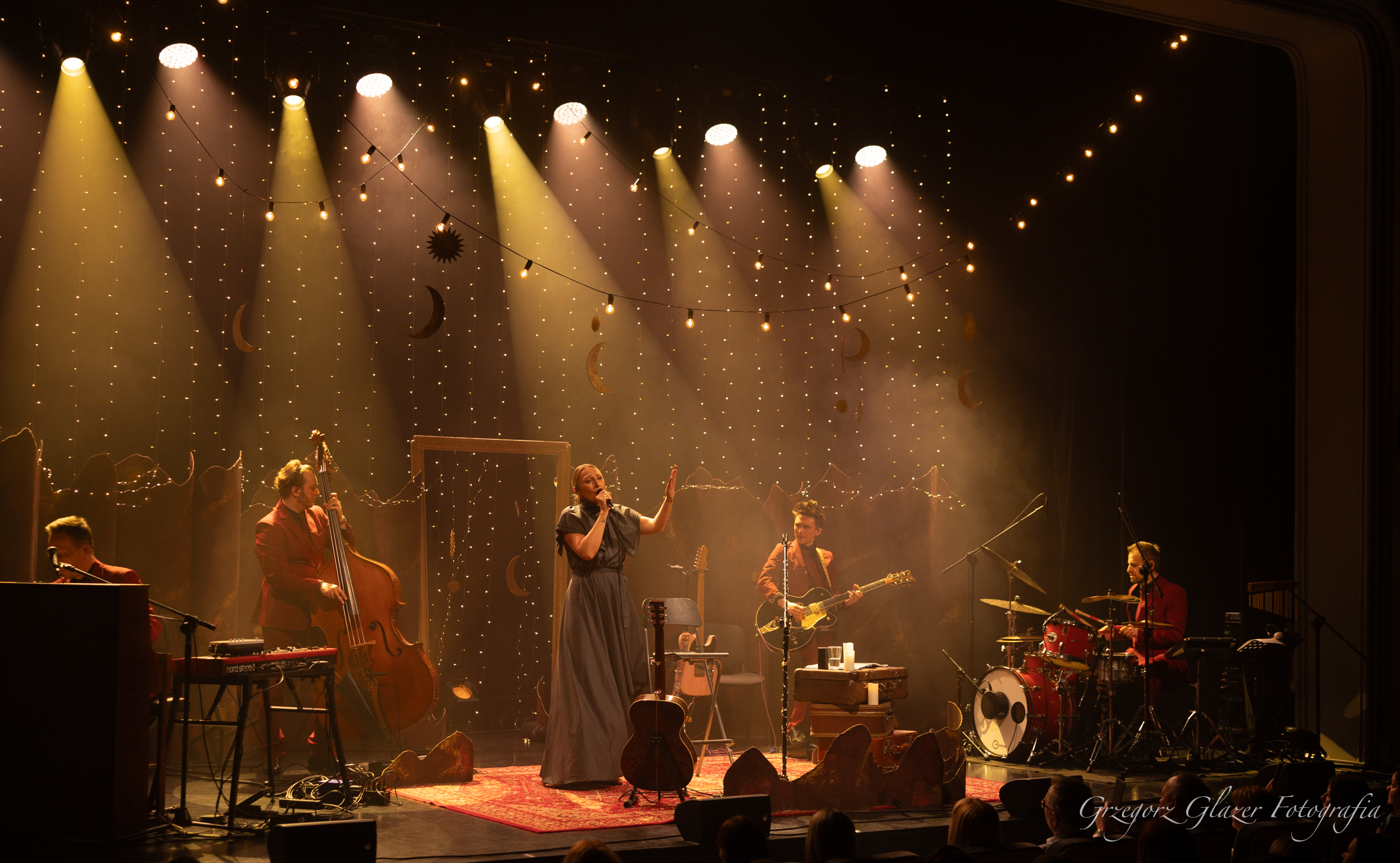 Kobieta o blond włosach siedzi na krześle i spiewa do mikrofonu, za nią na instrumentach grają męźczyżni w czerwonych garniturach.