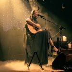 Kobieta o blond włosach i ciemnej sukience siedzi na krzesle, spiewa do mikrofonu i trzyma giatrę.