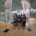 Grupa osób stoi w jasnym budynku, na podłodze z ułożonych płytek, za nimi znajdują się tory pływackie, po bokach stoją wysokie białe z napisami chorągiewki.