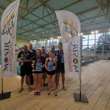 Grupa osób stoi w jasnym budynku pływalni i pozują do zdjęcia, za nimi tory pływackie.