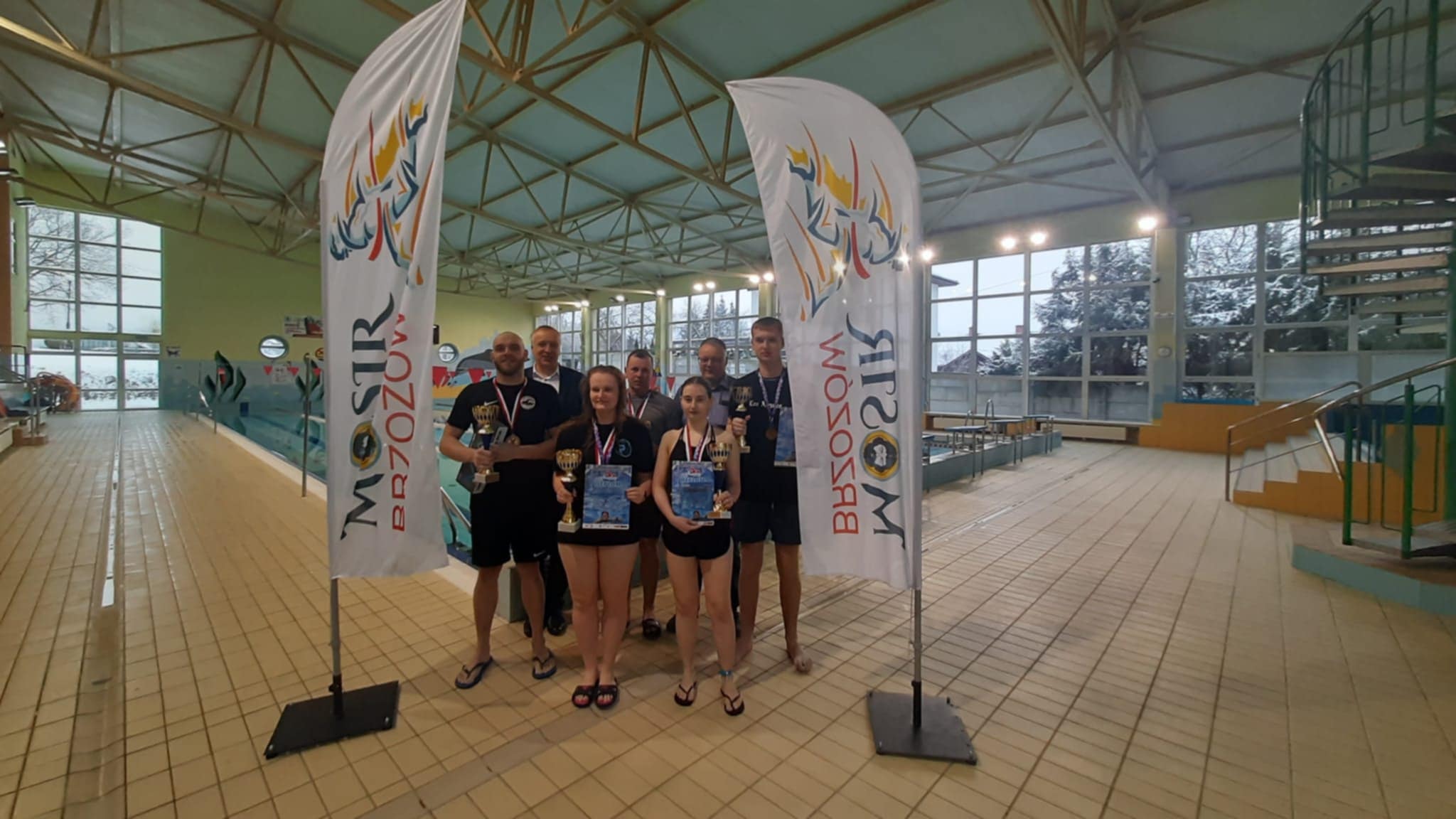 Grupa osób stoi w jasnym budynku pływalni i pozują do zdjęcia, za nimi tory pływackie.