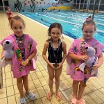 Dziewczynka ubrana w czarny stroj kąpielowy stoi wraz z dwiema dziewczynkami ubranymi w ręczniki różowe na basenie. Za nimi tory pływackie. Dziewczynki z różowymi ręcznikami trzymają w rękach maskotki.