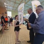 Młoda kobieta w ubrany krótki czarny strój odbiera medal i dyplom od dwóch starszych mężczyzn w garniturach. Wszyscy stoją na basenie.