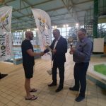 Mężczyzna w średnim wieku ubrany w krótki czarny strój odbiera medal i dyplom od dwóch starszych mężczyzn w garniturach. Wszyscy stoją na basenie.