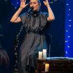 Kobieta o blond włosach w szarej sukience siedzi na krzesle i spiewa do mikrofonu.