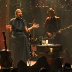 Kobieta o blond włosach i szarej sukience siedzi na krzesle na scenie i spiewa. Za nią stoi mężczyzna w garniturze i gra na gitarze.