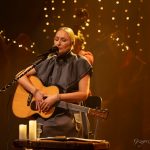 Kobieta o blond włosach i ciemnej sukience siedzi na krzesle, spiewa do mikrofonu i trzyma giatrę.