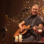 Kobieta o blond włosach i ciemnej sukience siedzi na krzesle, spiewa do mikrofonu i trzyma giatrę.