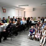 Grupa osób siedzi przy stole nakrytym białym obrusem, z boku siedzą na laweczkach dzieci ubrane w stroje ludowe.