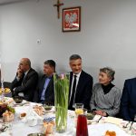 Grupa osób siedzi przy stole nakrytym białem obrusem.