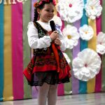 Dziewczynka w białej bluzce i sukience ludowej śpiewa do mikrofonu.