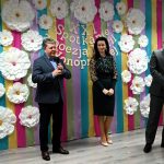 Mężczyzna w średnim wieku w garniturze, obok młoda kobieta o czarnych włosach oraz mężczyzna w garniturze w okularach przemawiają do mikrofonu.