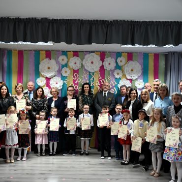 Grupa osób straszych i dzieci pozuje do wspólnego zdjęcia na tle ścianki wyłożonej kolorową bibułą.