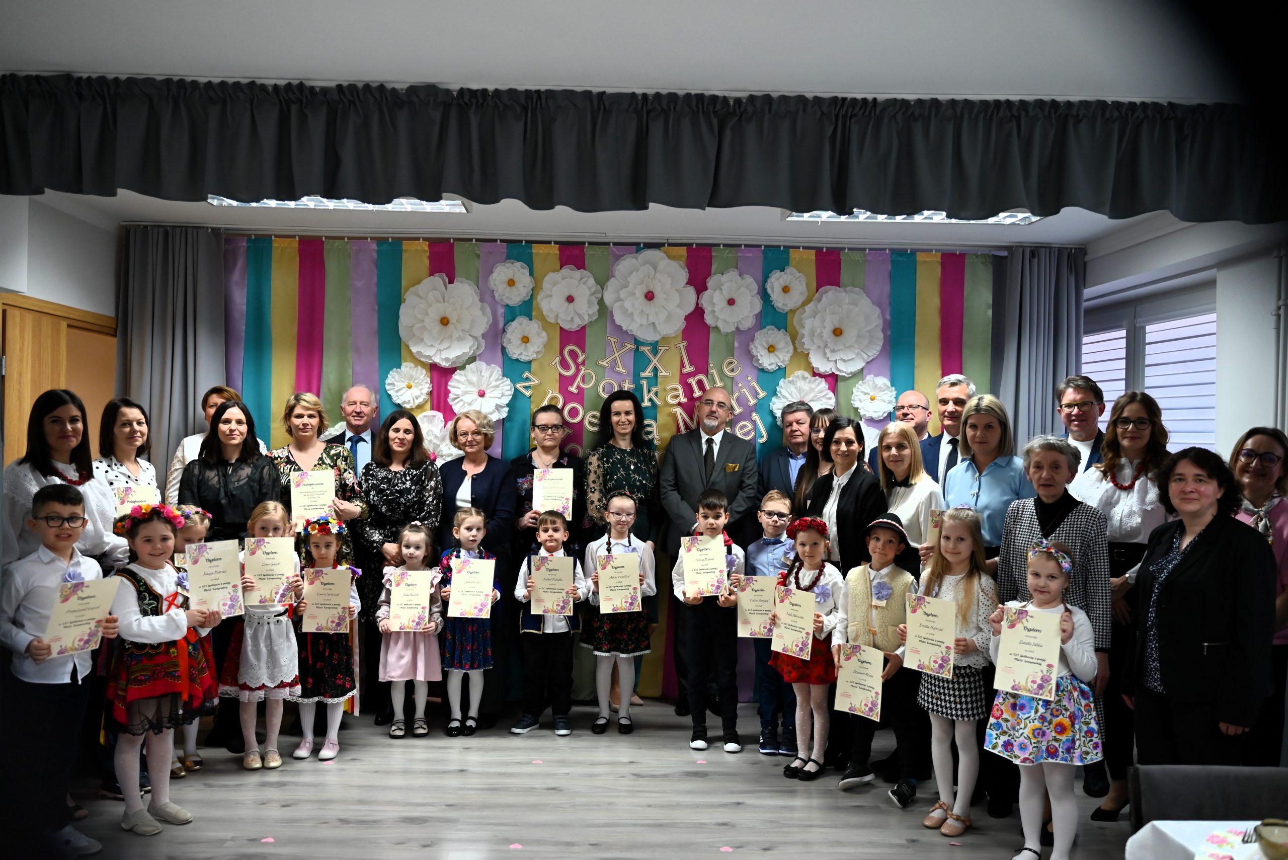 Grupa osób straszych i dzieci pozuje do wspólnego zdjęcia na tle ścianki wyłożonej kolorową bibułą.