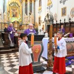 Wnętrze kościoła, starszy mężczyzna ubrany w fioletowy habiit przemawia do mikrofonu, przed mównicą stoją młodzi chłopcy i trzymają w rękach duże świece.