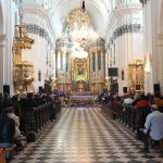 Wnętrze kościoła, na wprost duży pozłacony ołtarz, po bokach na ławkach siedzi kilkadziesiąt osób.