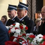 Starsi mężczyżni ubrani w galowe mundury, trzymają kwiaty biało-czerwone.