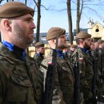 Mężczyźni ubrani w mundury wojskowe ze strzelbami stoją na placu przed kościołem.