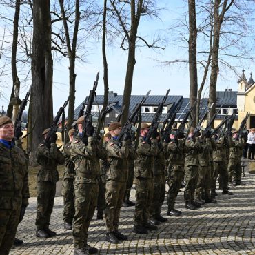 Mężczyźni ubrani w mundury wojskowe ze strzelbami stoją na placu przed kościołem.