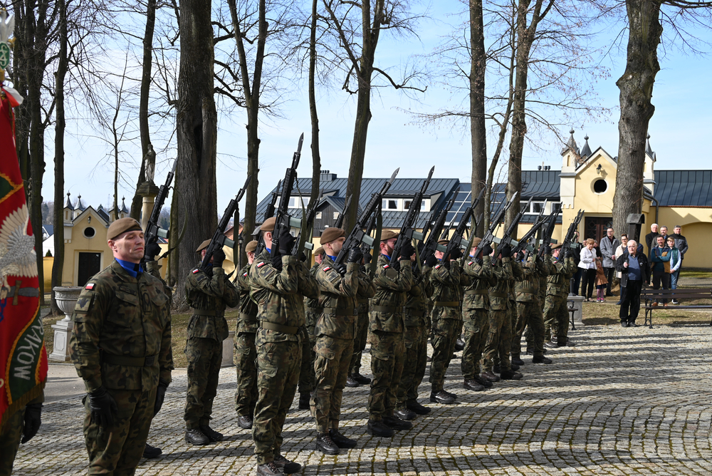 Mężczyźni ubrani w mundury wojskowe ze strzelbami stoją na placu przed kościołem.