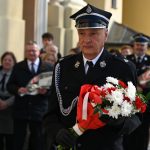 Mężczyzna ubrany w galowy mundur, trzyma kwiaty biało-czerwone.